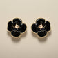 Black flower-shaped earrings with gold accents on a textured surface