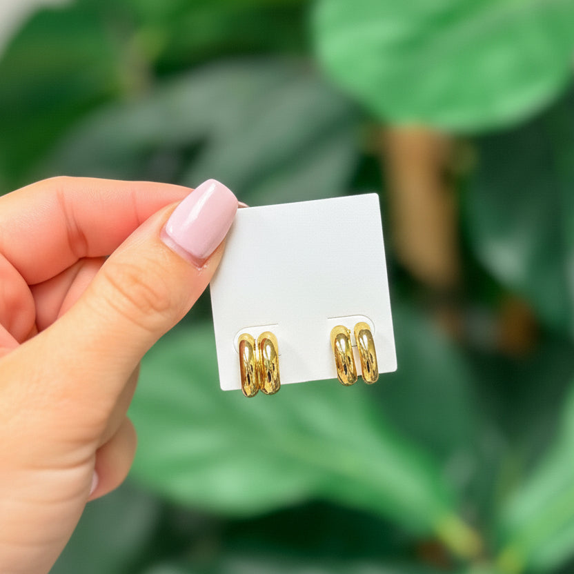 A hand holding a gold-dipped chunky huggie hoop earring with a pushback post, against a backdrop of green leaves.