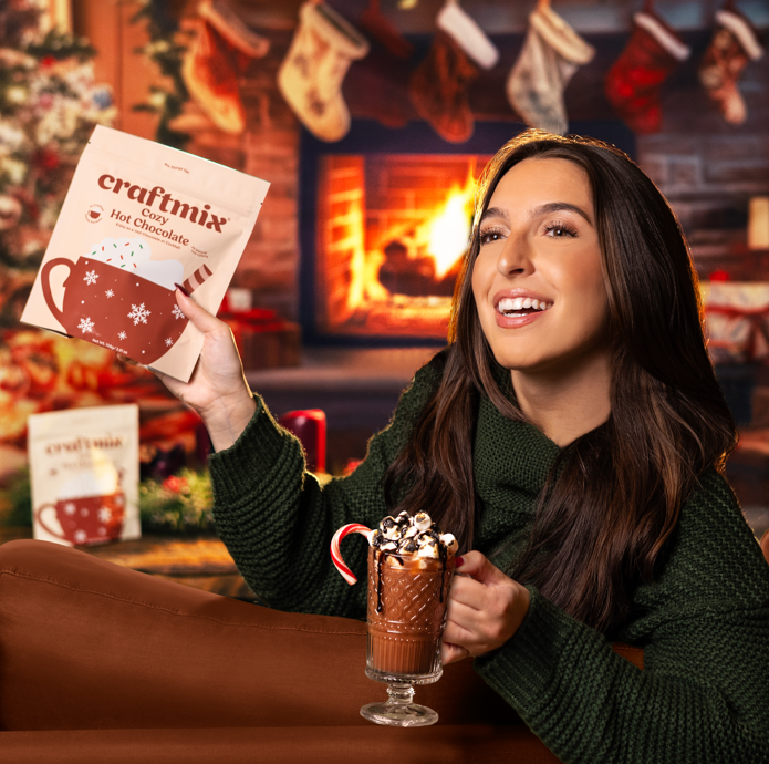Woman holding a craftmix hot chocolate packet and a mug of hot chocolate in a festive setting with stockings and a fireplace.