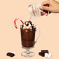 Hot chocolate mix being poured into a glass with a candy cane, surrounded by chocolate pieces and marshmallows on a beige background.