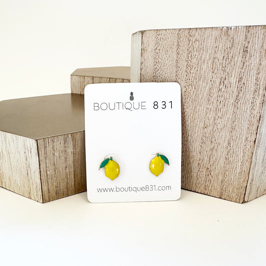 A pair of yellow lemon-shaped stud earrings displayed on a white card with 'Boutique 811' written on it, presented against a backdrop of wooden geometric shapes.