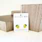 A pair of yellow lemon-shaped stud earrings displayed on a white card with 'Boutique 811' written on it, presented against a backdrop of wooden geometric shapes.