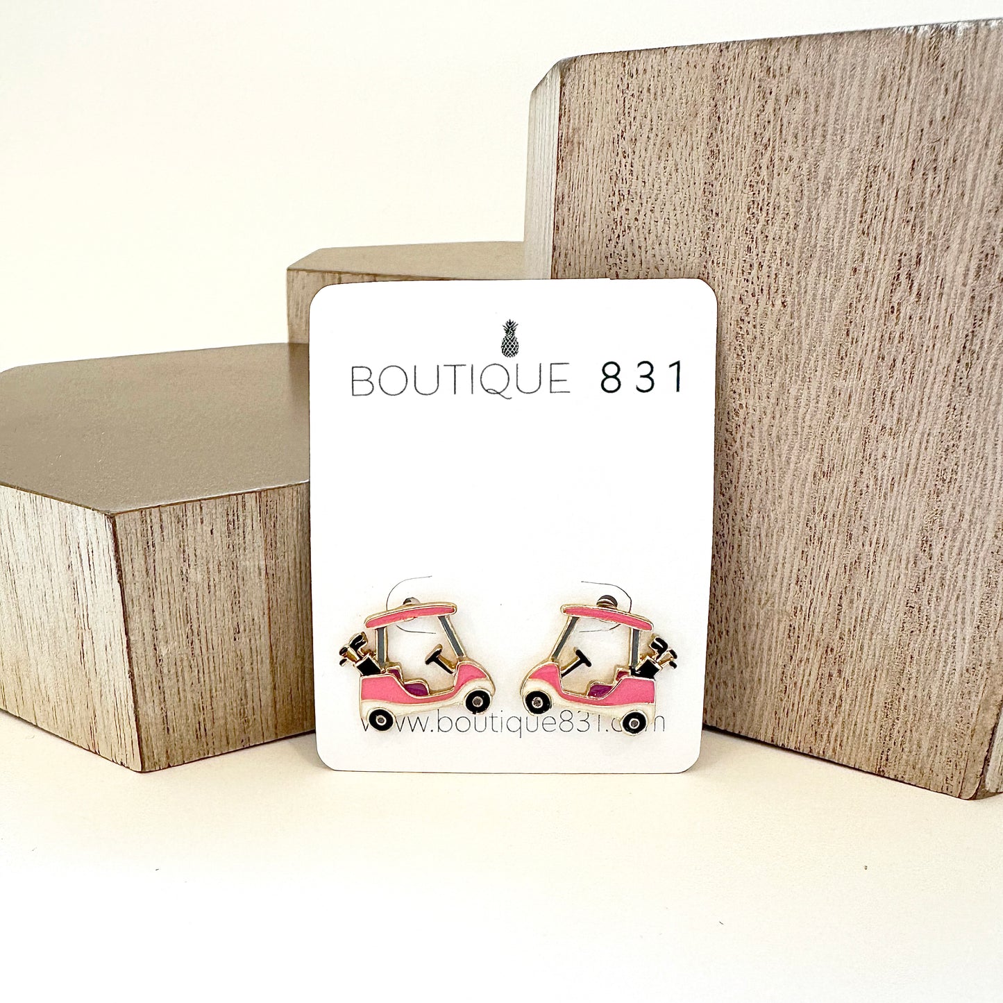 A pair of pink golf cart stud earrings against a geometric backdrop